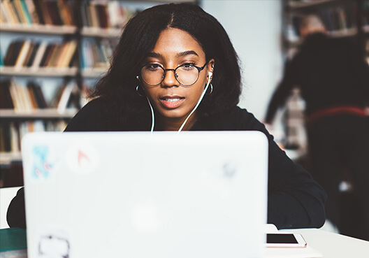 Young woman sitting in front of a bookshelf reading our policies on her laptop.